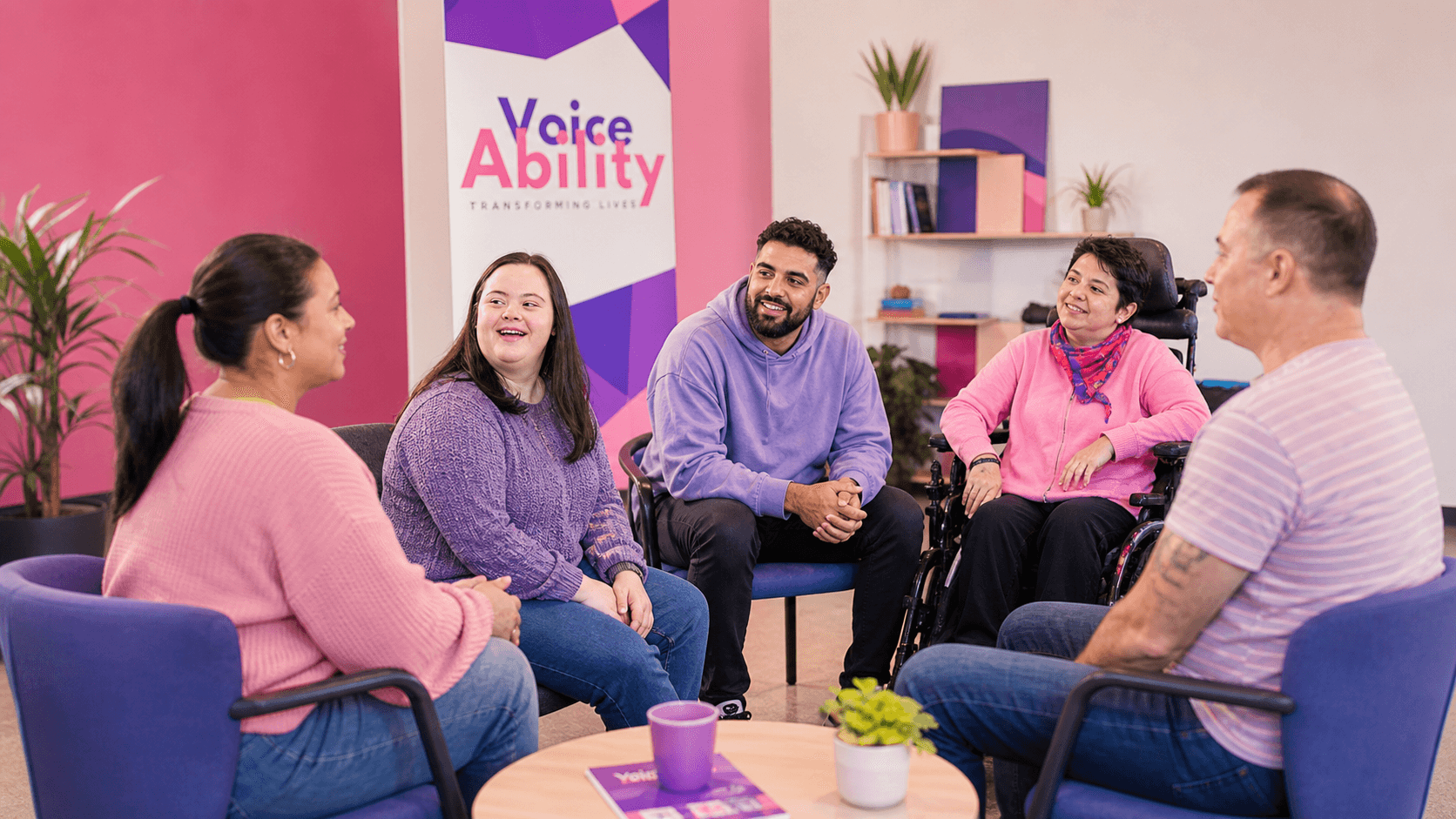 Five diverse people, including individuals with disabilities, sit in a circle and talk together.