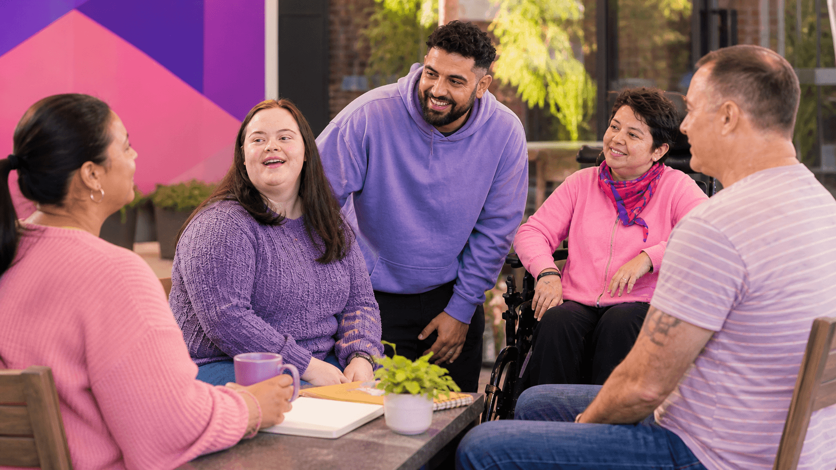 Diverse group of smiling people, including a woman in a wheelchair, talking around a table.