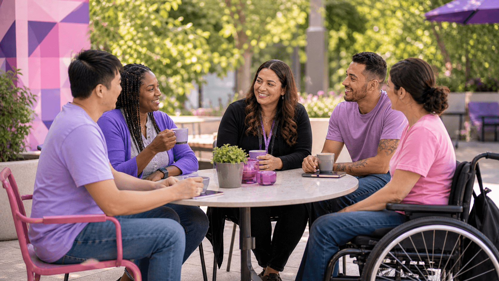 Five diverse people, including a wheelchair user, chatting over coffee at an outdoor table.
