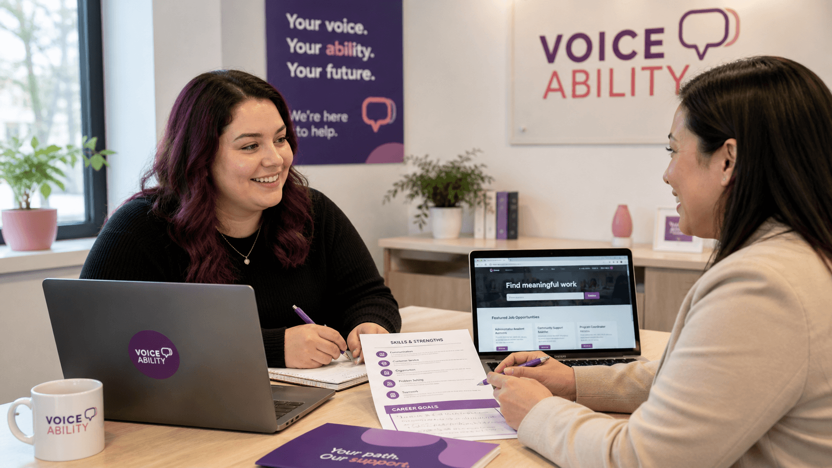 Two women smiling and discussing career goals with laptops and documents in a VoiceAbility office.