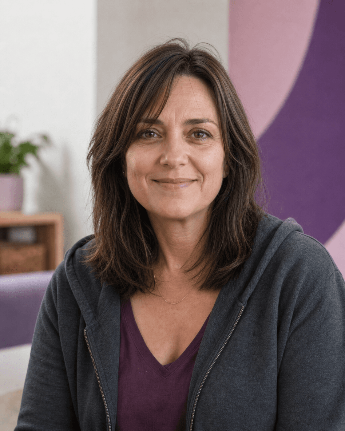 Smiling middle-aged woman with brown hair wearing a grey hoodie over a purple top.