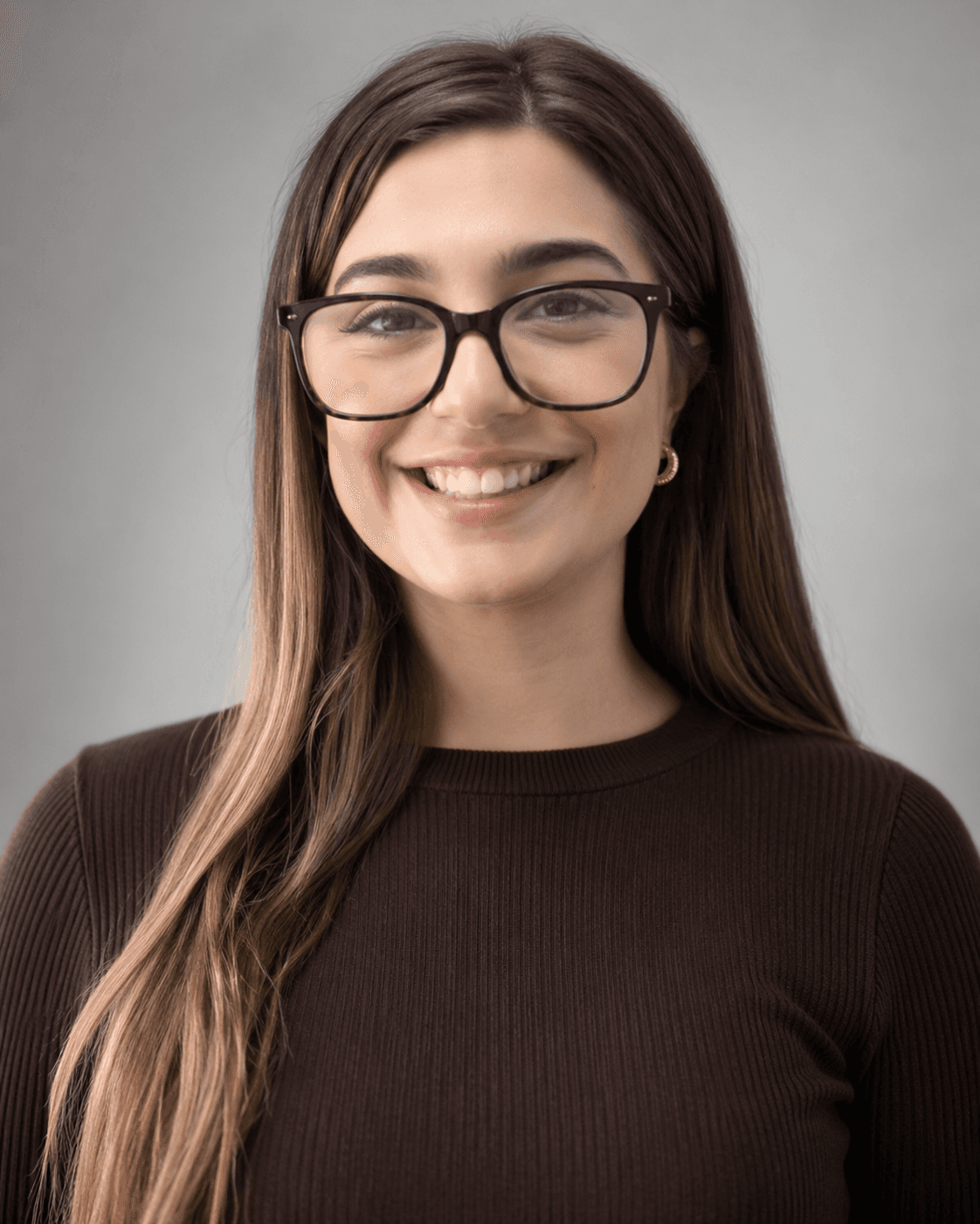 Smiling young woman with long brown hair, black-rimmed glasses, and a dark brown ribbed top.