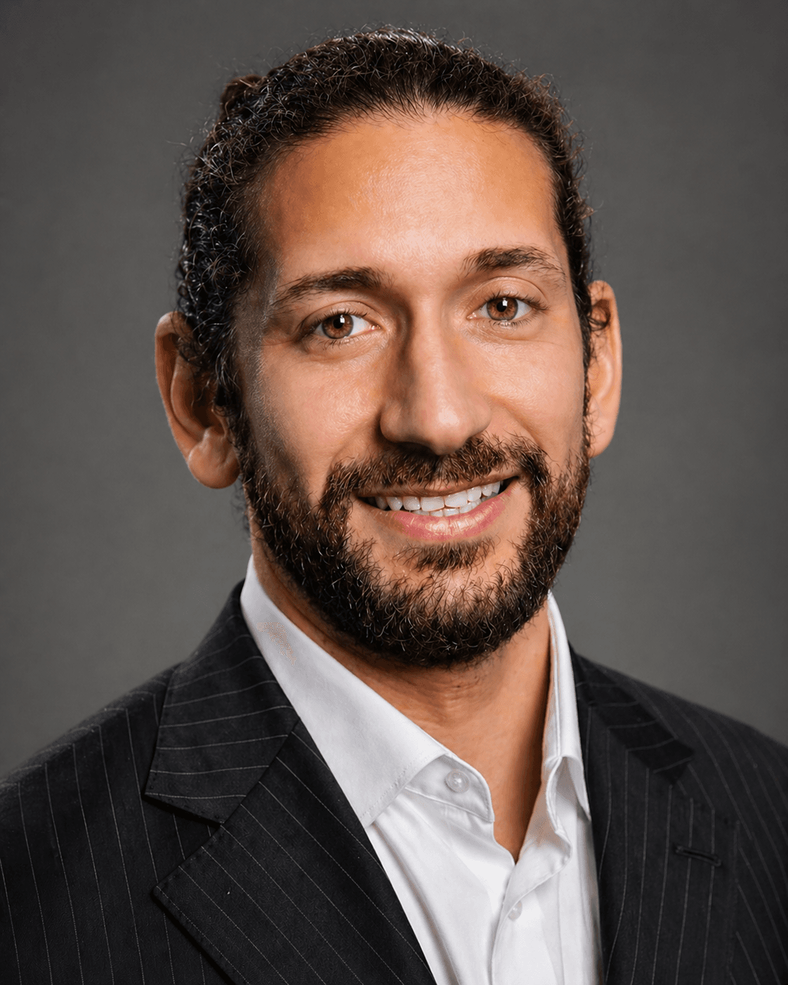 Smiling man with dark curly hair and a beard wearing a pinstriped suit jacket.