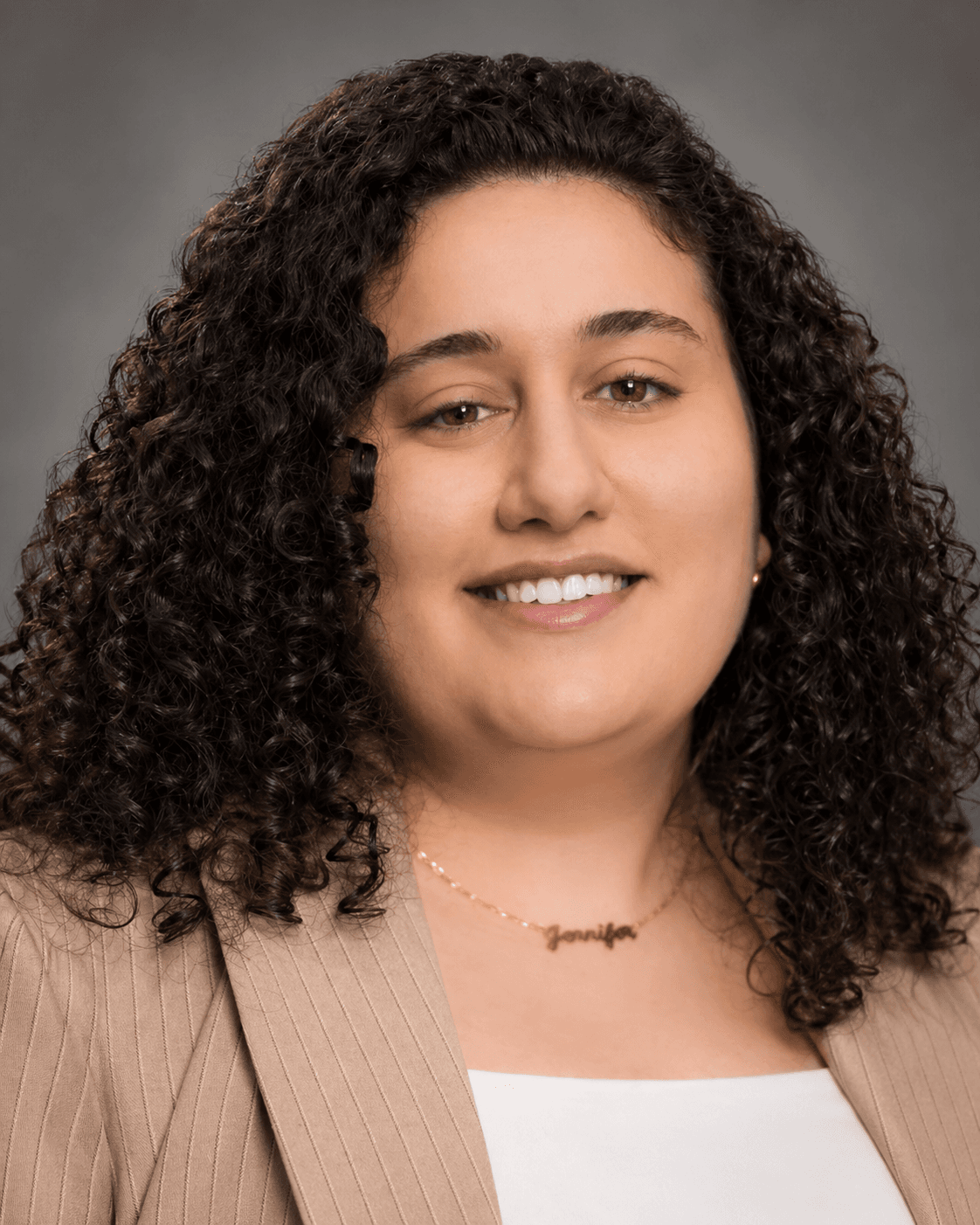 Smiling woman with dark curly hair wearing a tan pinstripe blazer and gold name necklace.